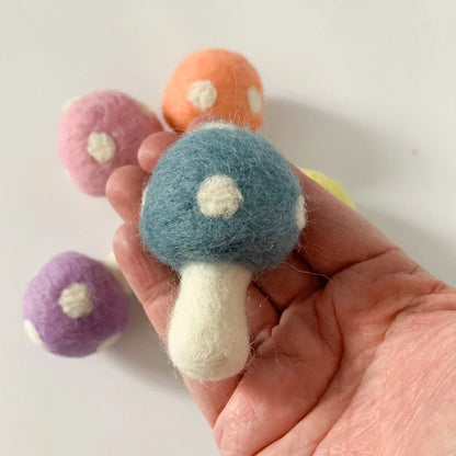 A hand holds a felted wool mushroom toy in blue and cream, with four more felted wool mushrooms in pink, orange, purple, and yellow blurred in the background against a plain white surface.
