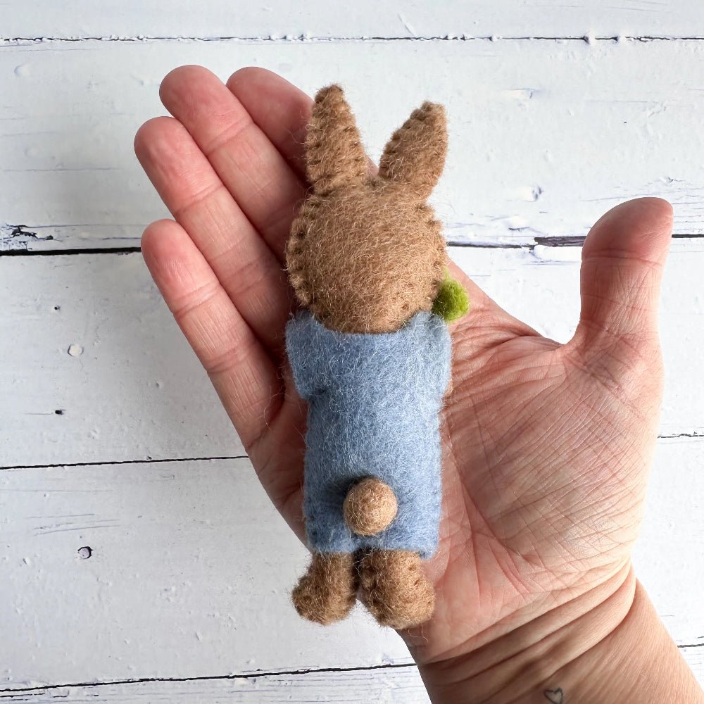 Hand holding a felted brown bunny, showcasing the back view against a white wooden background
