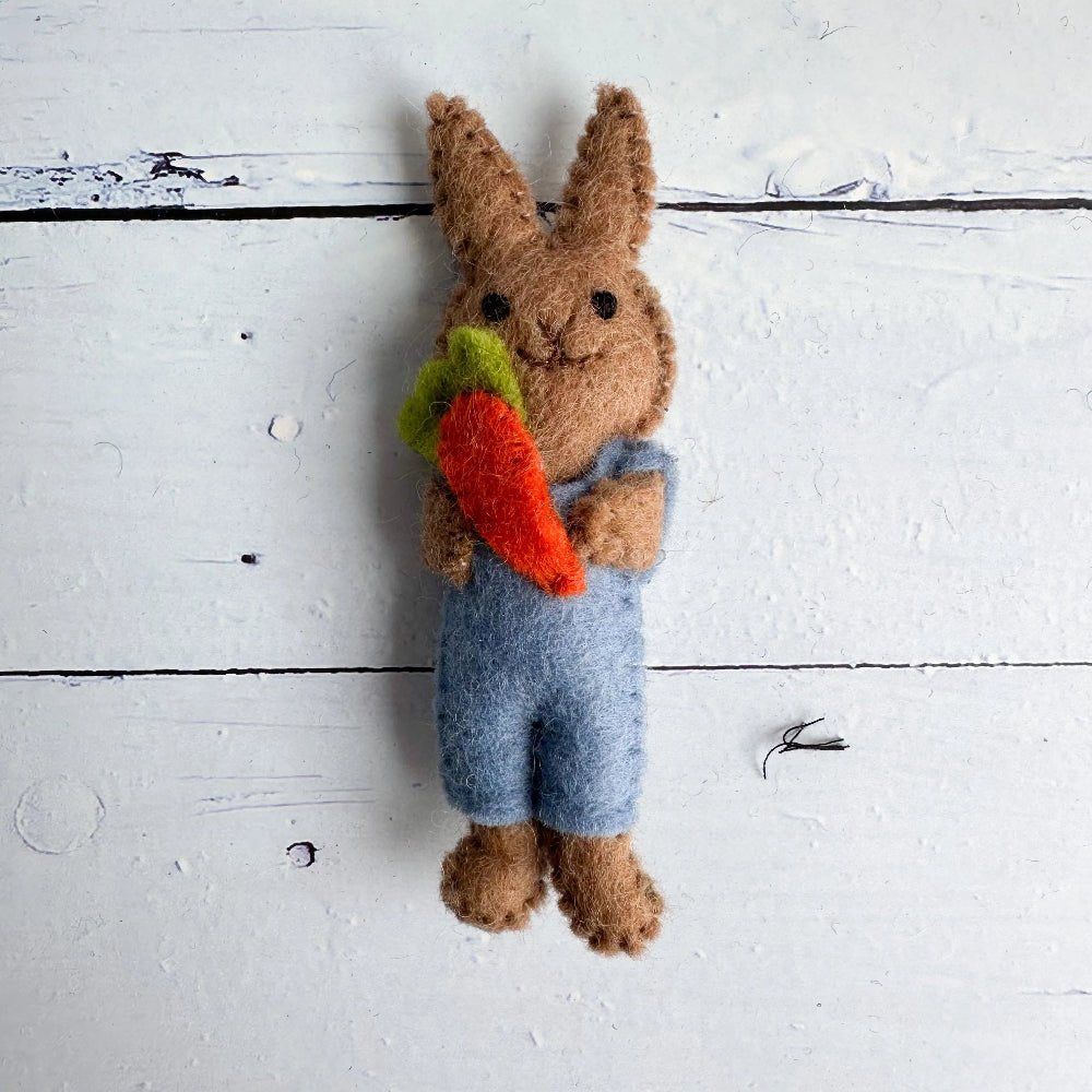 Felted bunny wearing blue overalls and holding a carrot on a wooden surface