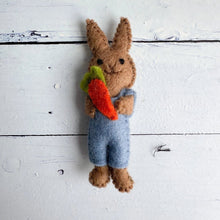 Felted bunny wearing blue overalls and holding a carrot on a wooden surface