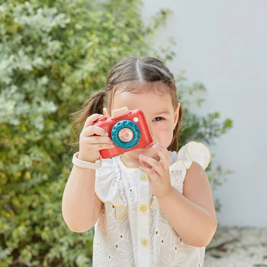 Young girl holding a red toy camera outdoors with greenery in the background