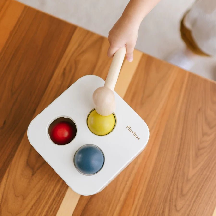 Child's hand holding a wooden mallet over a white toy box with colorful balls on a wooden floor.