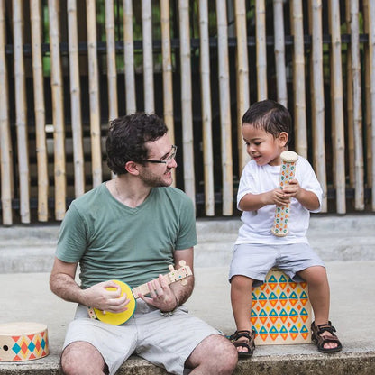 Adult playing a toy ukulele with a child holding a toy rain maker outdoors.