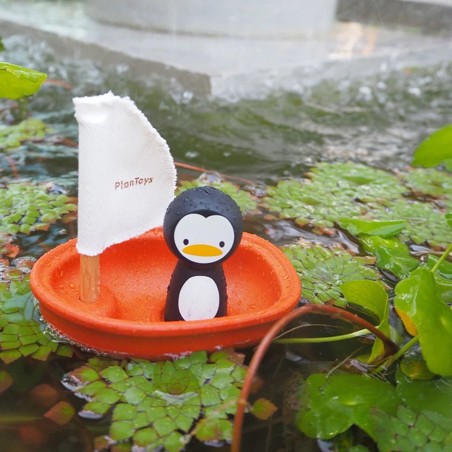 Toy penguin in an orange boat with a white sail, floating on water with green plants.