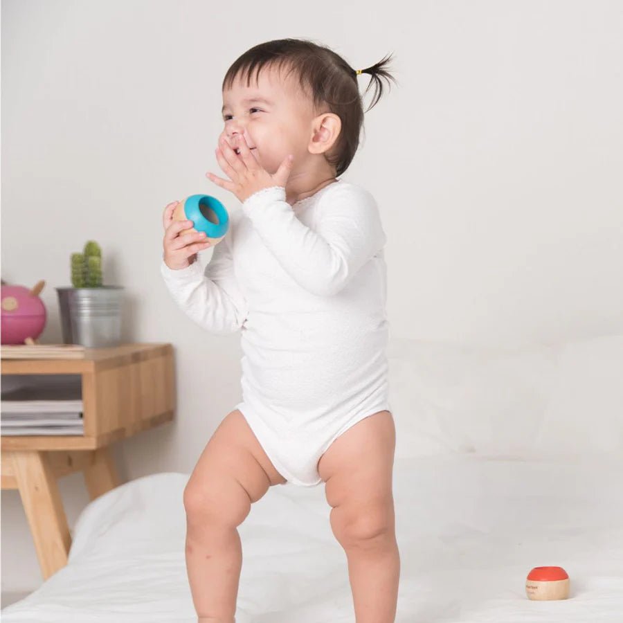 Child in a white onesie standing on a bed holding a blue sensory toy against a plain background