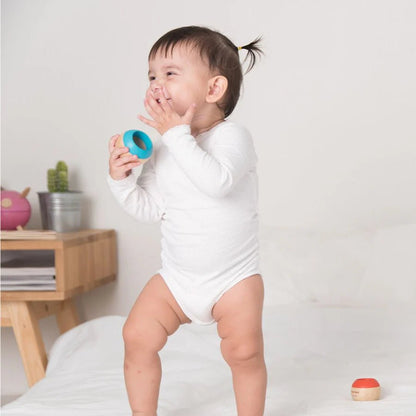 Child in a white onesie standing on a bed holding a blue sensory toy against a plain background
