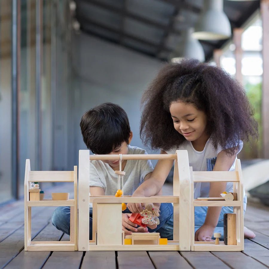 Two children playing with a wooden doll house on a wooden deck.