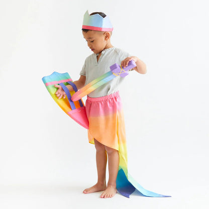 Child wearing a rainbow-colored mermaid tail and a crown, holding a sword and a shield, on a white background