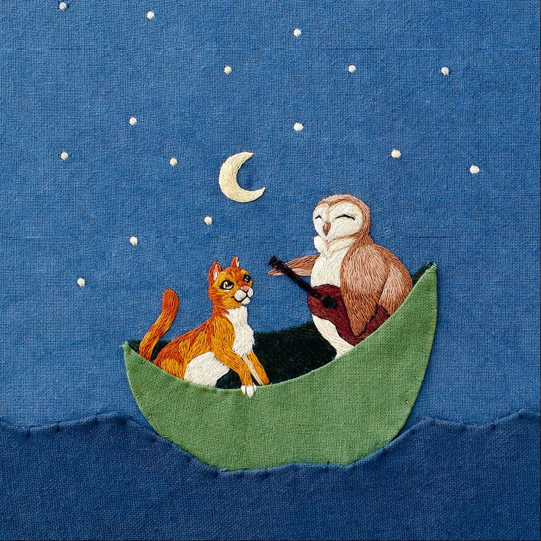 Embroidered image of an owl playing guitar to an orange cat in a boat on the water