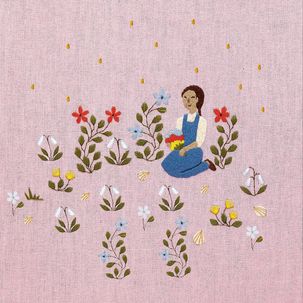 Embroidered artwork featuring a girl sitting in a field of flowers on a pink background