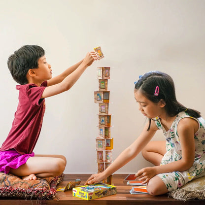 Two children playing with a card tower on a wooden floor.