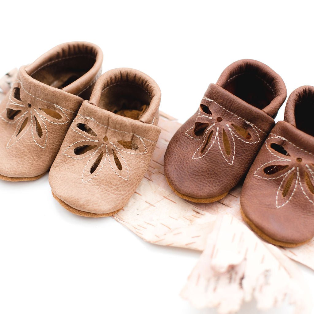 Four baby moccasin-style shoes arranged on a white surface with a pale pink fringed textile. Two shoes are light tan/beige and two are medium brown, each with a floral cutout detail on the top and white contrast stitching.