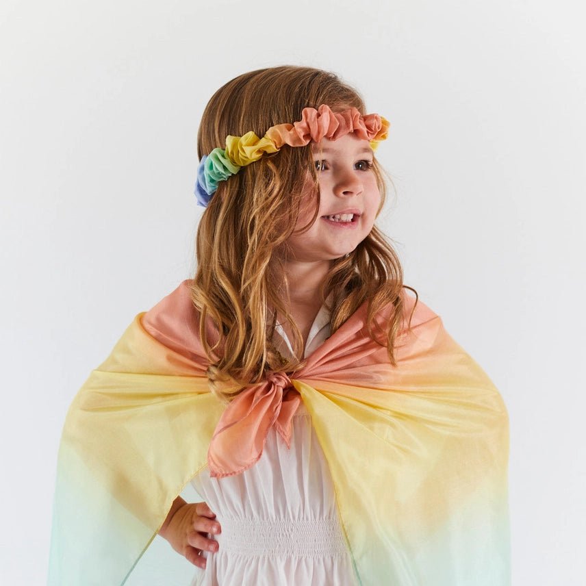 Young girl wearing a pastel rainbow-colored garland and cape on a white background