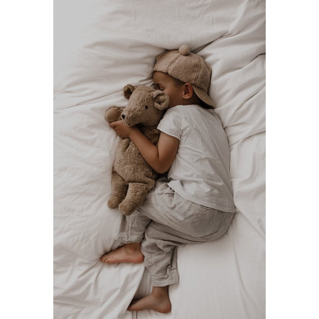 Child lying in bed with a Senger cuddly teddy bear, wearing a matching cap and outfit.