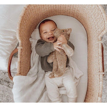Baby holding a Senger beige teddy bear  in a wicker crib with a soft, neutral background
