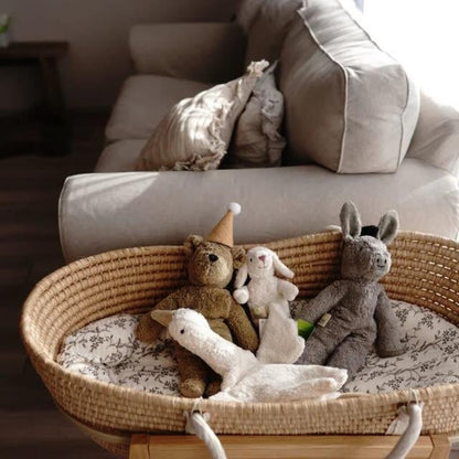 Wicker basket with stuffed animals includeing a Senger beige teddy bear  on a floral-patterned cushion in front of a beige sofa.