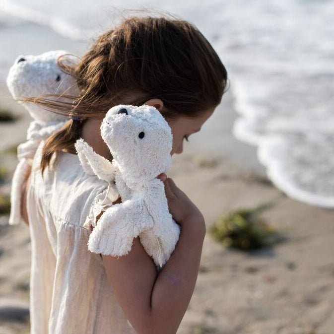 Child holding two plush white seals on a beach with ocean waves in the background