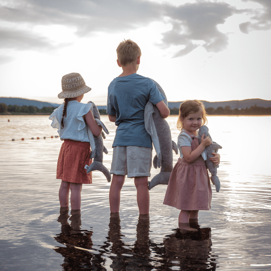 Three children standing in water, looking at a lake with mountains in the background while holding shark stuffed animals