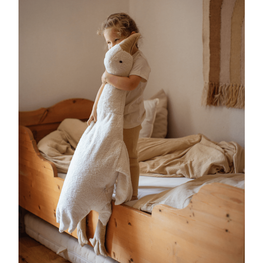Child holding a large white stuffed animal goose on a wooden bed in a cozy room.