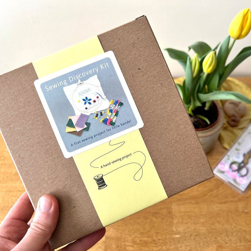 Sewing discovery kit with a hand holding it, placed on a wooden surface with a plant in the background.