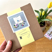 Sewing discovery kit with a hand holding it, placed on a wooden surface with a plant in the background.