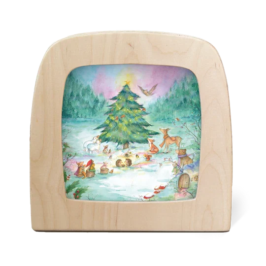 A square wooden picture frame with rounded corners, featuring a colorful painted winter forest scene with a Christmas tree, animals, and a snowy ground, shown on a plain white background.