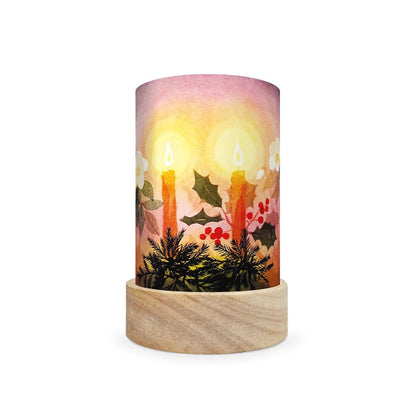 A cylindrical table lamp shade with a light natural wood base and a printed wraparound design featuring two glowing candles, holly leaves, berries, and a soft pink-to-orange sunset background.