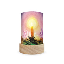 A cylindrical table lamp shade with a light wood base and a printed cylinder body featuring a watercolor-style candle glowing in front of crystals under a pink and purple sky.