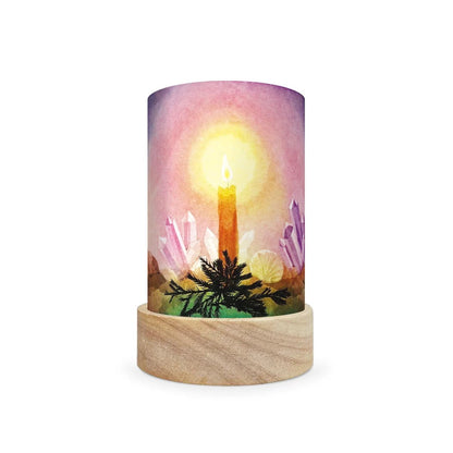 A cylindrical table lamp shade with a light wood base and a printed cylinder body featuring a watercolor-style candle glowing in front of crystals under a pink and purple sky.