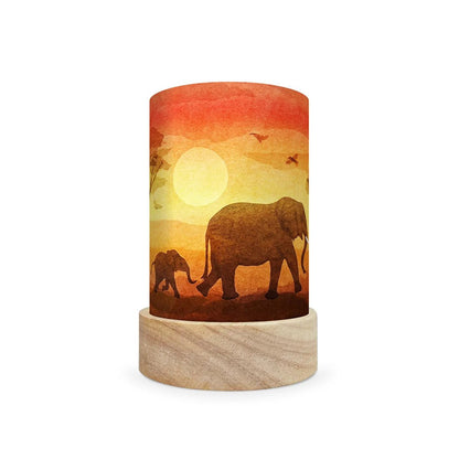 A cylindrical decorative lamp shade with a printed sunset scene featuring elephants and a large sun, mounted on a light wood base against a white background.