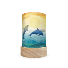 A cylindrical decorative lamp shade with a light natural wood base and a printed wrap featuring two dolphins over blue water under a yellow sun, shown on a plain white background.