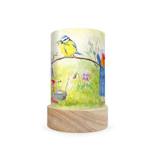 A cylindrical table lamp with a light wood base and a printed wraparound cylinder featuring a watercolor-style illustration of a bluebird perched on a branch above a grassy garden scene, shown on a plain white background.