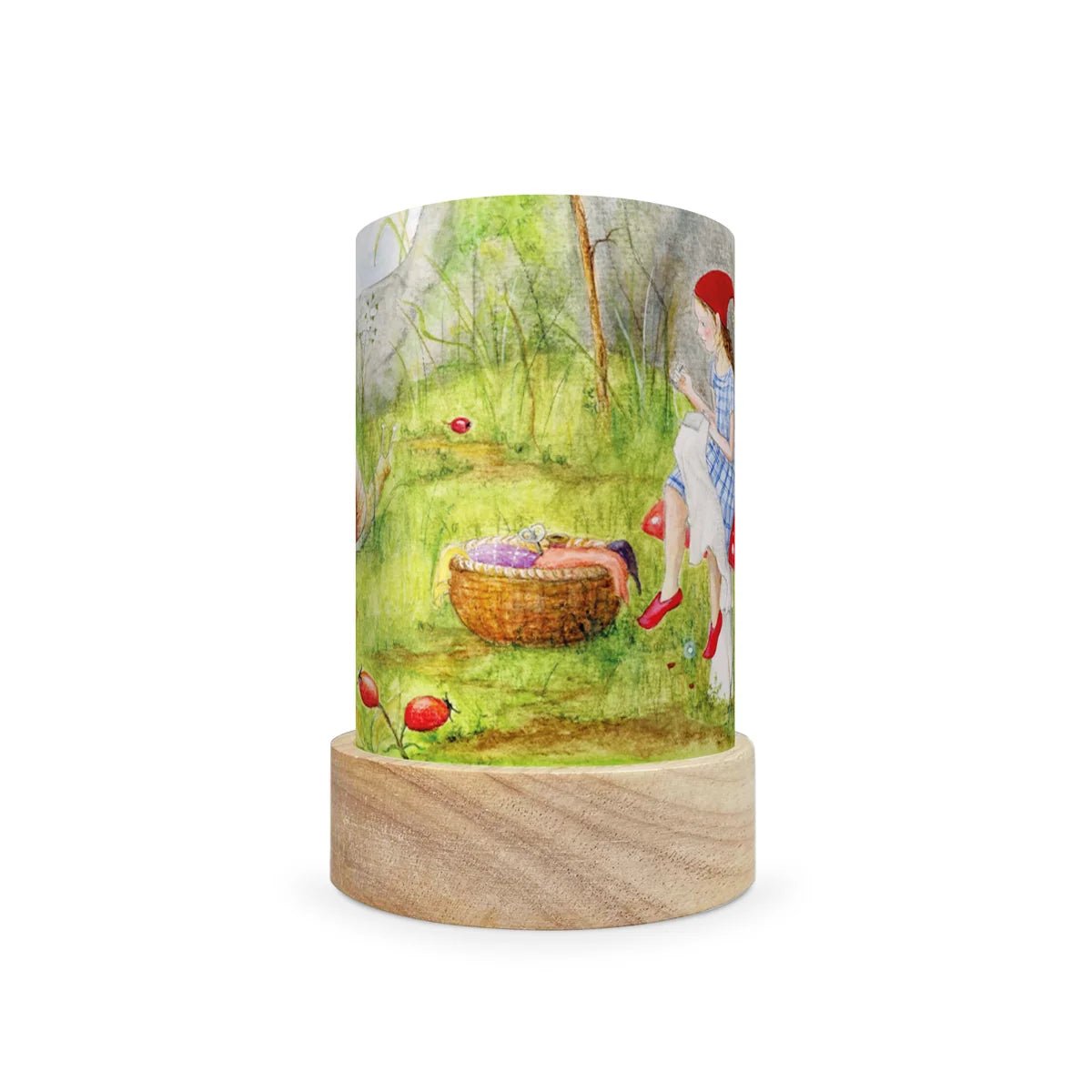 A cylindrical table lamp with a light wood base and a printed fabric shade featuring a watercolor-style illustration of a girl in a red hat and dress near a basket in a green outdoor scene.