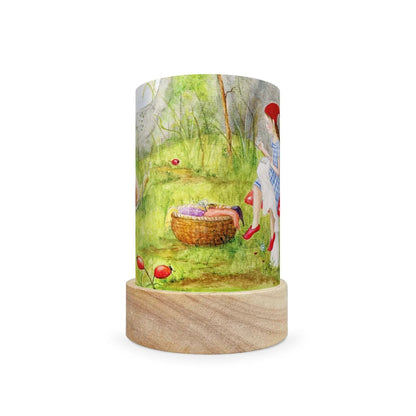 A cylindrical table lamp with a light wood base and a printed fabric shade featuring a watercolor-style illustration of a girl in a red hat and dress near a basket in a green outdoor scene.