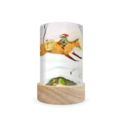 A cylindrical table lamp with a light wood base and a printed wraparound cylinder featuring an illustration of a fox carrying a rider through a snowy landscape, shown on a plain white background.