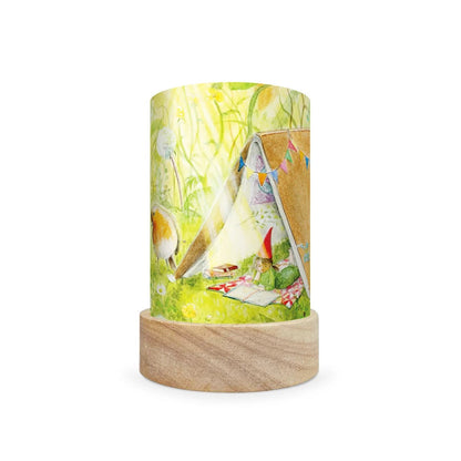 A cylindrical table lamp with a light wood base and a printed wraparound cylinder featuring a whimsical camping scene with a small tent, green grass, and a gnome, shown on a white background.