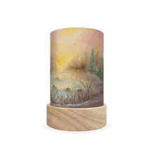 Decorative lamp with a wooden base and painted scene  of christmas trees on the edge of a lake on a white background