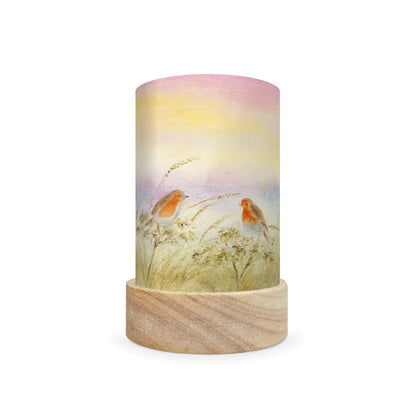 A cylindrical decorative lamp shade with a light wood base and a printed wraparound cylinder featuring a pastel sunset sky and two small birds in a grassy field.