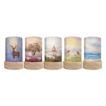 Five cylindrical decorative lamp shades with printed nature-themed artwork, shown in a row against a plain white background. The designs include a stag, a tree in winter, a woman in a field, birds in grass, and an ocean scene.