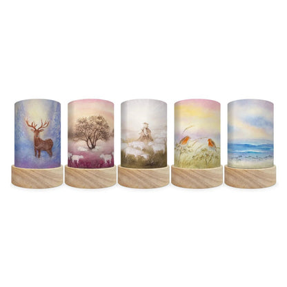 Five cylindrical decorative lamp shades with printed nature-themed artwork, shown in a row against a plain white background. The designs include a stag, a tree in winter, a woman in a field, birds in grass, and an ocean scene.