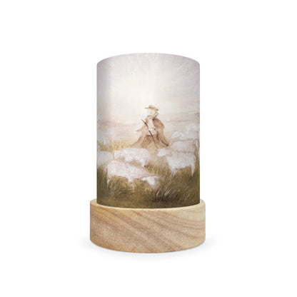 A cylindrical decorative lamp shade with a light wood base and a printed cylinder shade featuring a soft, vintage-style illustration of a shepherd with a staff among sheep in a misty field.
