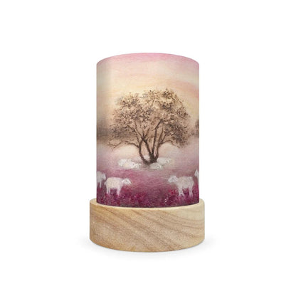 A cylindrical decorative lamp shade with a light wood base and a printed cylinder body featuring a scenic landscape of a tree and sheep in a field under a pink sky.