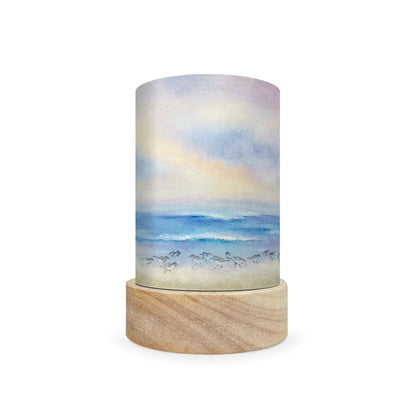 A cylindrical decorative lamp shade with a watercolor-style ocean scene printed on the shade, resting on a round natural wood base against a plain white background.