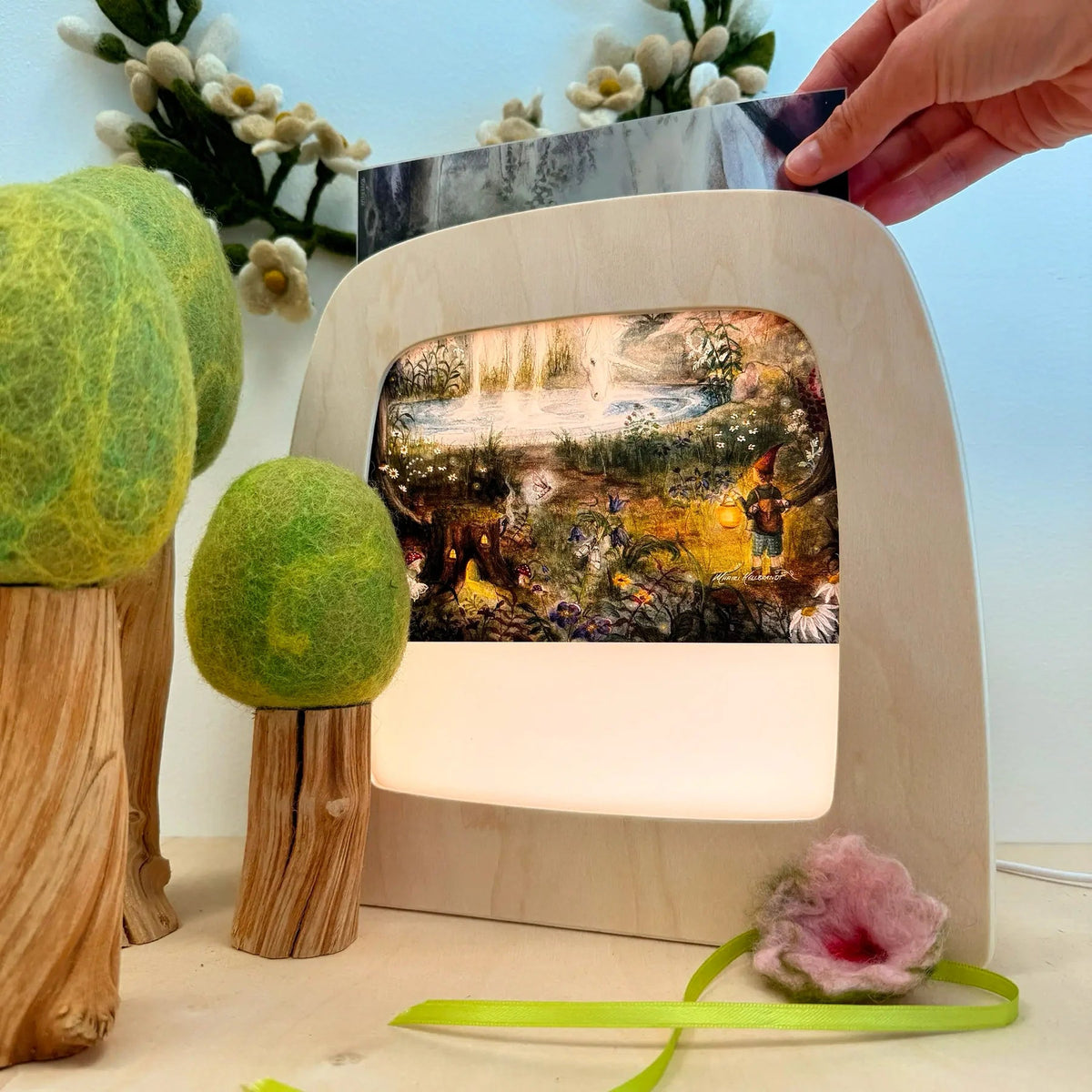 A hand holds a silhouette above a Toverlux lamp that glows with a colorful forest scene. The setup is styled with felt trees and flowers on a tabletop against a pale blue background.