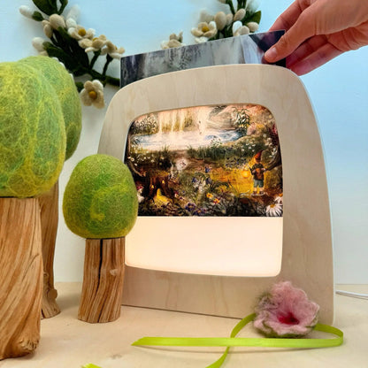 A hand holds a silhouette above a Toverlux lamp that glows with a colorful forest scene. The setup is styled with felt trees and flowers on a tabletop against a pale blue background.