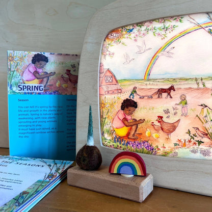 Through the seasons EduLux book nex to Spring silhouette depicting a Black girl feeding chickens, with a rainbow in the background