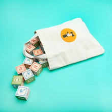 Colorful wooden blocks spilling out of a white drawstring bag with a logo on a teal background