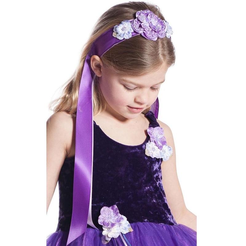 A young girl wearing a purple flower girl costume dress with a matching purple headband adorned with purple and white fabric flowers, looking down against a white background.
