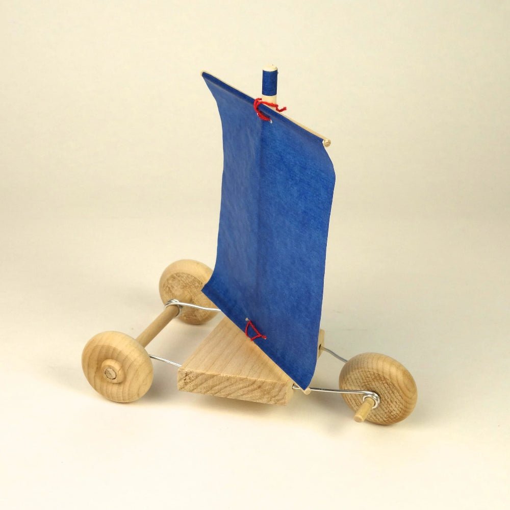 A small wooden sailing cart with a blue fabric sail, natural wood wheels, and a simple metal wire frame, photographed on a plain light beige studio background.