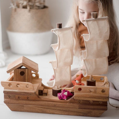 A young girl with long blonde hair sits on a white floor next to a wooden toy pirate ship with beige canvas sails, in a bright, softly lit room.